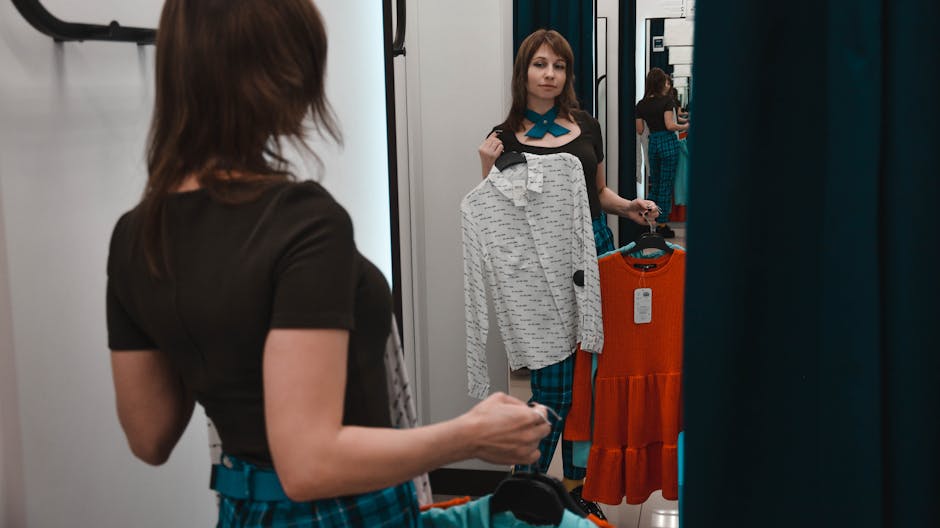 Back view of young woman standing in front of mirror in changing room of store and picking shirts
